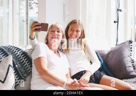 Lächelnde Enkelin nimmt Selfie mit Großmutter über Handy auf Sofa zu Hause Stockfoto