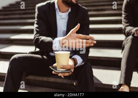 Mittelteil des Geschäftsmannes mit Einweg-Tasse macht Hand Geste, während Sitzen auf der Treppe Stockfoto