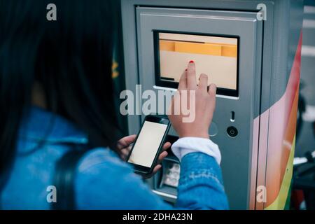 Rückansicht eines Teenagers, der den Geldautomaten berührt, während er hält Smartphone Stockfoto