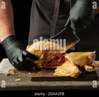 Koch in schwarzer Uniform und Latexhandschuhe hacken rohes Huhn In Stücke auf einem braunen Holzbrett Stockfoto