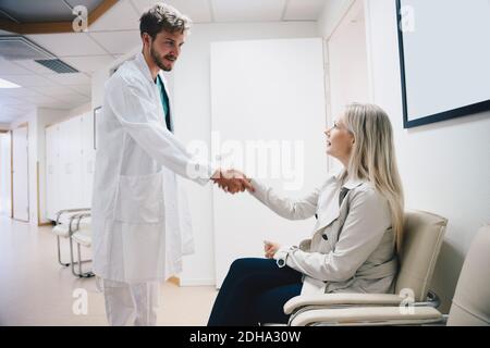 Junger männlicher Arzt begrüßt weibliche Patienten auf Stuhl sitzen in Krankenhausflur Stockfoto