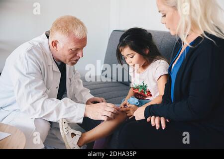Reifen männlichen Arzt Prüfung Mädchen Knie, während von der Mutter sitzen Auf dem Sofa im Krankenhaus Stockfoto