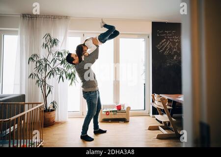 Voller Länge des verspielten Vaters hebt seine Tochter im Stehen Im Wohnzimmer Stockfoto