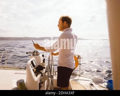 Seitenansicht des Mannes, der während des Segelns des Katamarans das Mobiltelefon hält Auf dem Meer gegen den Himmel während des sonnigen Tages Stockfoto