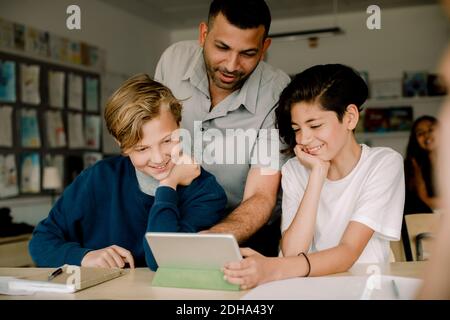 Lächelnder männlicher Tutor mit Schülern, die während des Sitzens ein digitales Tablet benutzen Im Klassenzimmer Stockfoto