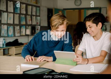 Lächelnde männliche Schüler, die im Klassenzimmer mit einem digitalen Tablet sitzen Stockfoto