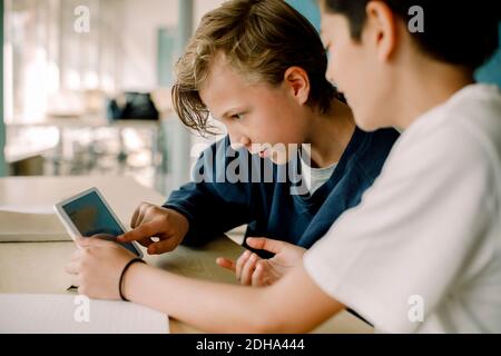 Männlicher Student zeigt auf digitale Tablette, während er mit einem Freund sitzt Im Klassenzimmer Stockfoto