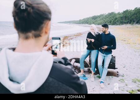 Rückansicht eines jungen Mannes, der Freunde fotografiert, die auf Metall sitzen Am Strand Stockfoto
