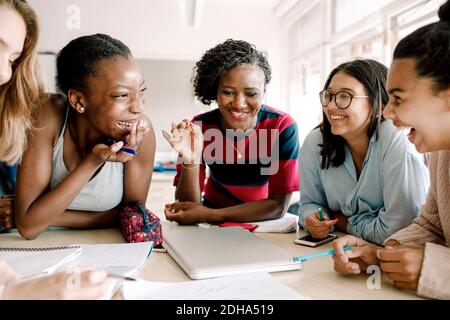 Lächelnder Lehrer gestikulierte, während fröhliche Schüler am Tisch sitzen Schulungsraum Stockfoto