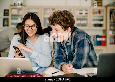 Lächelndes Teenager-Mädchen zeigt Smartphone zu männlichen Freund, während Sitzen im Klassenzimmer Stockfoto