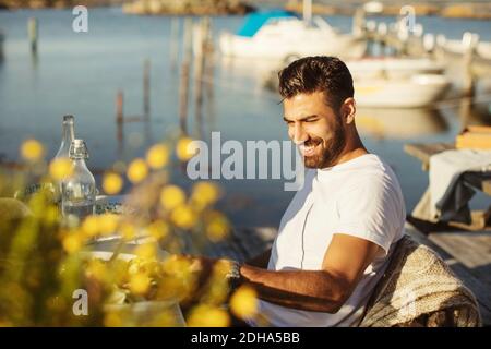 Lächelnder junger Mann, der auf einem Stuhl am Tisch gegen den See sitzt Stockfoto