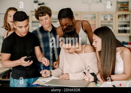 Lächelnder Teenager beim Lernen, während Freunde am Tisch im Klassenzimmer stehen Stockfoto