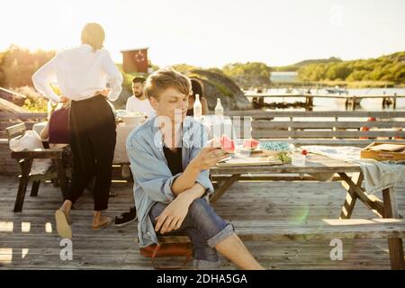 Lächelnder Mann beim Essen Wassermelone Scheibe, während Freunde im Hintergrund sitzen An der Anlegestelle Stockfoto