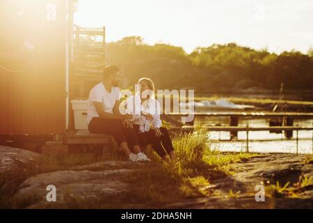 Volle Länge des glücklichen jungen Paares reden, während sitzen an Blockhütte am sonnigen Tag Stockfoto