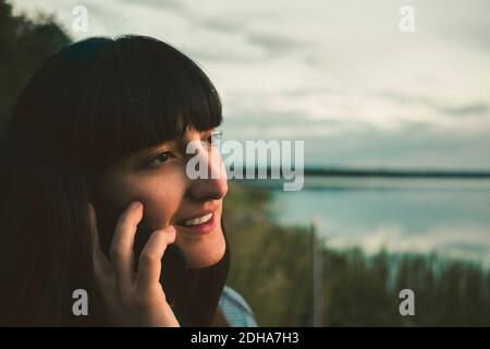 Lächelnde Frau, die am See gegen den Himmel auf dem Handy spricht Stockfoto