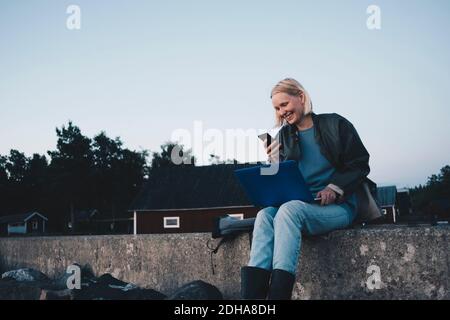 Lächelnde Frau, die auf das Mobiltelefon schaut und dabei den Laptop hält Sitzen auf Stützwand Stockfoto
