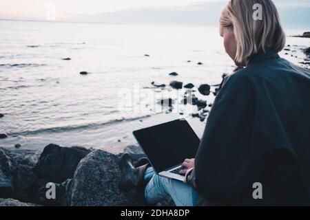 Rückansicht einer Frau, die einen Laptop benutzt, während sie auf Felsen sitzt Am Seeufer Stockfoto