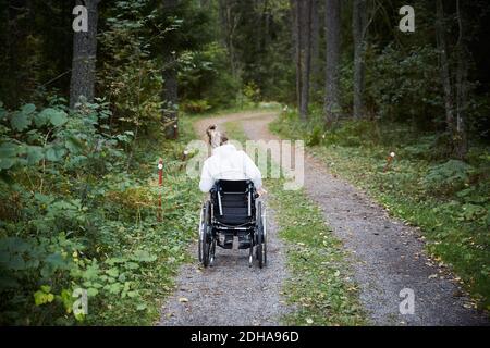 Rückansicht einer behinderten Frau im Rollstuhl auf unbefestigten Straßen Im Wald Stockfoto