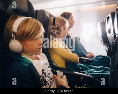 Kaukasischer Junge mit Kopfhörern sitzt mit Schwester und Mutter in Flugzeug Stockfoto