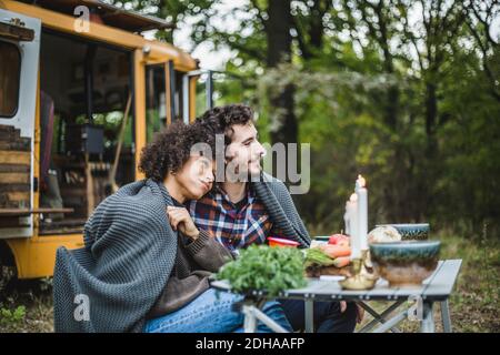 Junge multi-ethnische Paar in Decke gewickelt, während am Tisch sitzen Und wegschauen Stockfoto