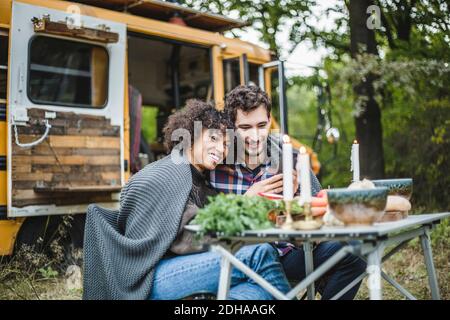Lächelndes junges Paar, das sich das Handy teilt, während es in eine Decke gehüllt ist Zusammen beim Camping im Wald Stockfoto