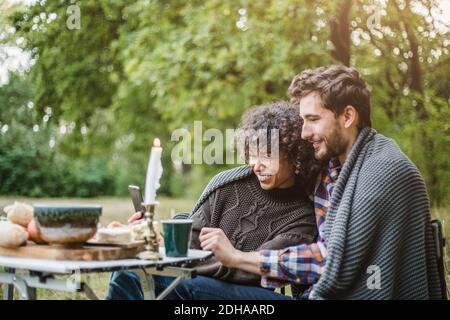 Fröhliches junges Paar, das Handy teilt, während es in eine Decke gehüllt ist Zusammen beim Camping im Wald Stockfoto