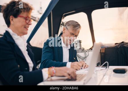 Älterer Mann, der auf Papier schreibt, während er mit einer Frau sitzt Laptop in der Yacht Stockfoto