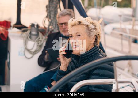 Ältere Frau, die auf dem Handy spricht, während sie bei einem Mann sitzt In Yacht Stockfoto