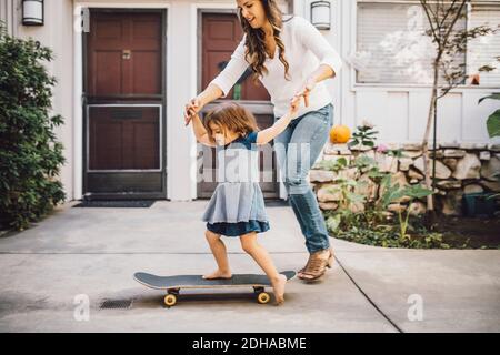 Lächelnde Tochter balanciert über Skateboard mit Hilfe von Mutter auf Fußweg Stockfoto