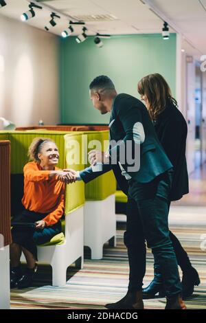 Lächelnde Geschäftsfrau sitzend, während sie die Hände schüttelt mit Geschäftsmann von Frau Kollege, der im Flur im Büro steht Stockfoto