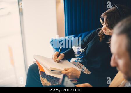 Junge Geschäftsfrau, die Kopfhörer hört, während sie auf dem Notizblock sitzt Von einem männlichen Kollegen in der Bürokantine Stockfoto