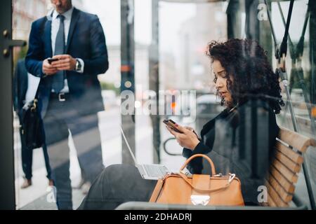 Geschäftsleute, die Smartphones benutzen, während sie in der Busstation warten Vom Glas aus gesehen Stockfoto