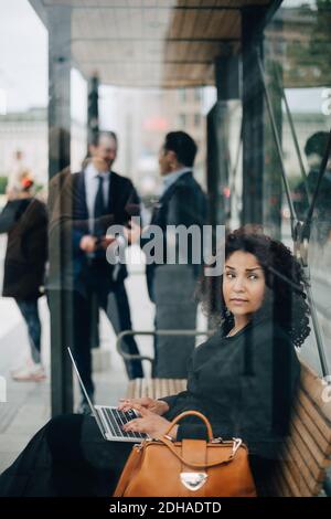 Seitenansicht einer Geschäftsfrau mittleren Erwachsenen, die beim Sitzen wegschaut Mit Laptop im Bus-Unterstand aus Glas gesehen Stockfoto