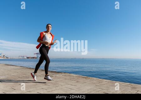 Seitenansicht der athletischen jungen Frau, die entlang der Strandpromenade läuft. Sportlerin beim Joggen am Meer Stockfoto