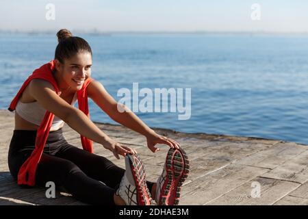 Außenaufnahme eines lächelnden, selbstbewussten Sportlertrainings in der Nähe des Meeres. Fitness-Frau sitzt auf hölzernen Pier und Stretching Beine Stockfoto
