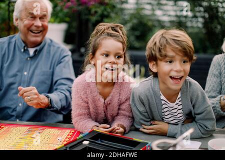 Lächelnde Enkelkinder spielen Brettspiel mit Großeltern auf dem Tisch in Hinterhof Stockfoto