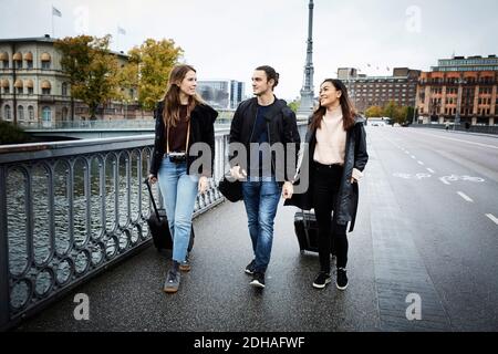 Männliche und weibliche Freunde sprechen beim Gehen mit Gepäck auf Brücke in der Stadt Stockfoto