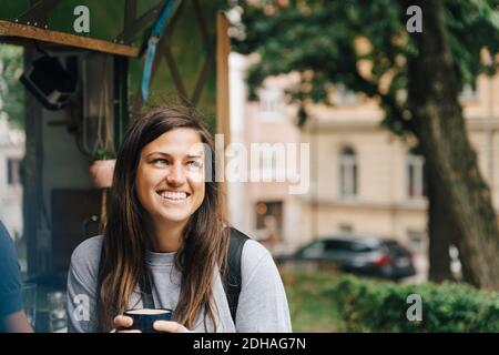 Lächelnde Frau hält Kaffeetasse, während sie am Konzessionsständer sitzt In der Stadt Stockfoto