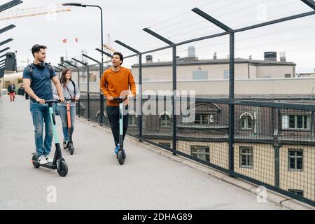 Volle Länge der Freunde mit Push-Scooter auf Brücke in Stadt gegen Himmel Stockfoto