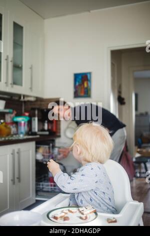 Blonde Baby Junge sitzt auf Hochstuhl beim Blick auf Vater in der Küche Stockfoto