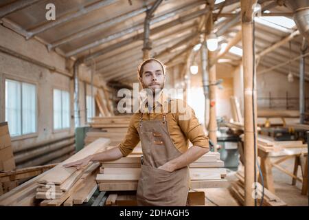 Porträt eines fröhlichen Zimmermanns, der neben dem rohen Holz steht Materialien im Lager der Schreinerei Stockfoto