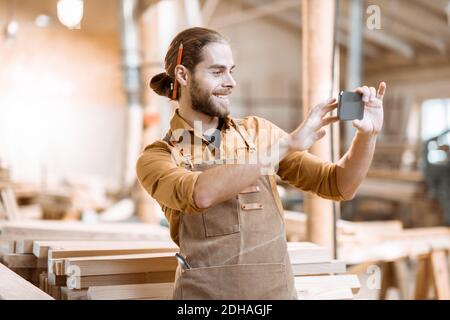 Schöner Zimmermann im Gespräch auf Smartphone oder mit dem Telefon, um einige Holzarbeiten in der Schreinerei zu entwerfen. Konzept der mobilen Technologien in der Zimmerei Stockfoto