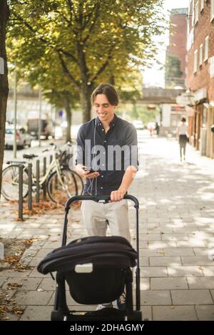 Lächelnder Mann, der Smartphone benutzt, während er den Kinderwagen anschiebt Fußweg in der Stadt Stockfoto