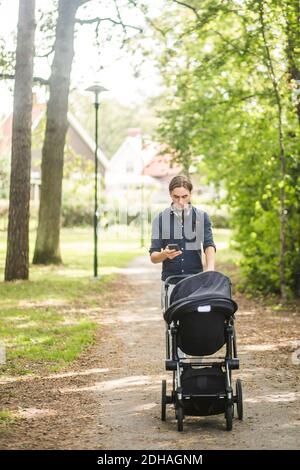 Mann, der Smartphone benutzt, während der Babywagen auf den Fußweg geschoben wird Im öffentlichen Park Stockfoto