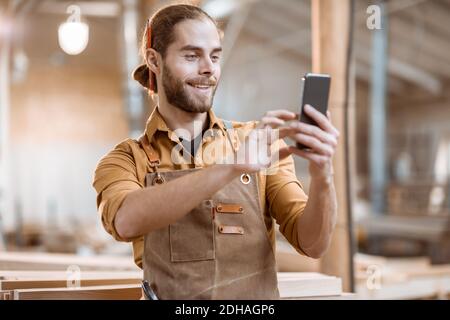 Schöner Zimmermann im Gespräch auf Smartphone oder mit dem Telefon, um einige Holzarbeiten in der Schreinerei zu entwerfen. Konzept der mobilen Technologien in der Zimmerei Stockfoto