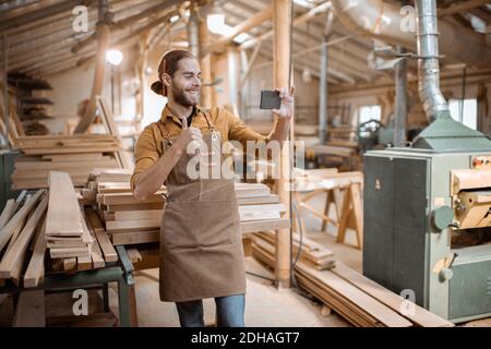 Schöner Zimmermann im Gespräch auf Smartphone oder mit dem Telefon, um einige Holzarbeiten in der Schreinerei zu entwerfen. Konzept der mobilen Technologien in der Zimmerei Stockfoto