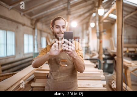 Schöner Zimmermann im Gespräch auf Smartphone oder mit dem Telefon, um einige Holzarbeiten in der Schreinerei zu entwerfen. Konzept der mobilen Technologien in der Zimmerei Stockfoto