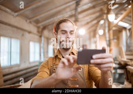 Schöner Zimmermann im Gespräch auf Smartphone oder mit dem Telefon, um einige Holzarbeiten in der Schreinerei zu entwerfen. Konzept der mobilen Technologien in der Zimmerei Stockfoto