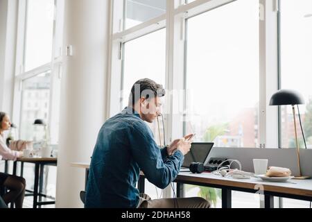 Junger Geschäftsmann mit Smartphone, während er mit Laptop sitzt Schreibtisch im Büro Stockfoto