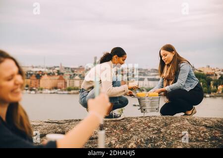 Weibliche Freunde rösten Zuckermais auf Felsformation durch See während Picknick Stockfoto